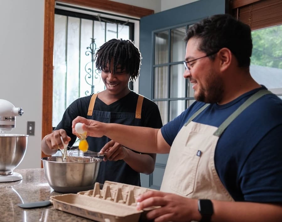 Two men people together, smiling. Younger cracks egg, older holds carton.